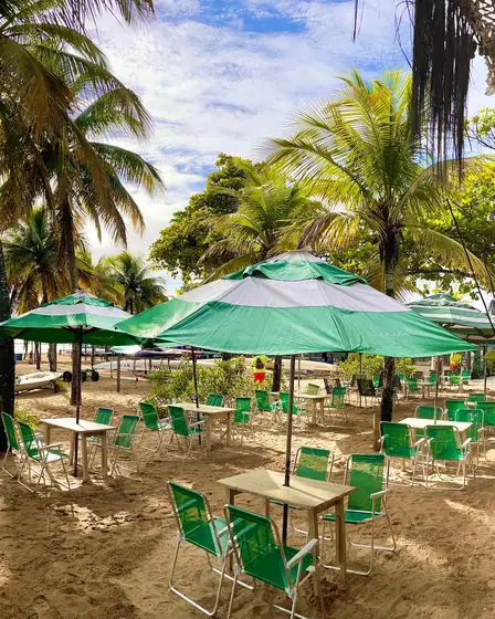 Mesas e cadeiras verdes na areia sob guarda-sóis listrados e coqueiros